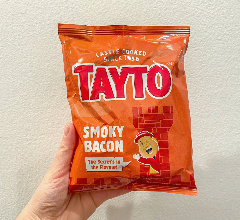 An orange bag of Tayto Brand Smoky Bacon Crisps being held