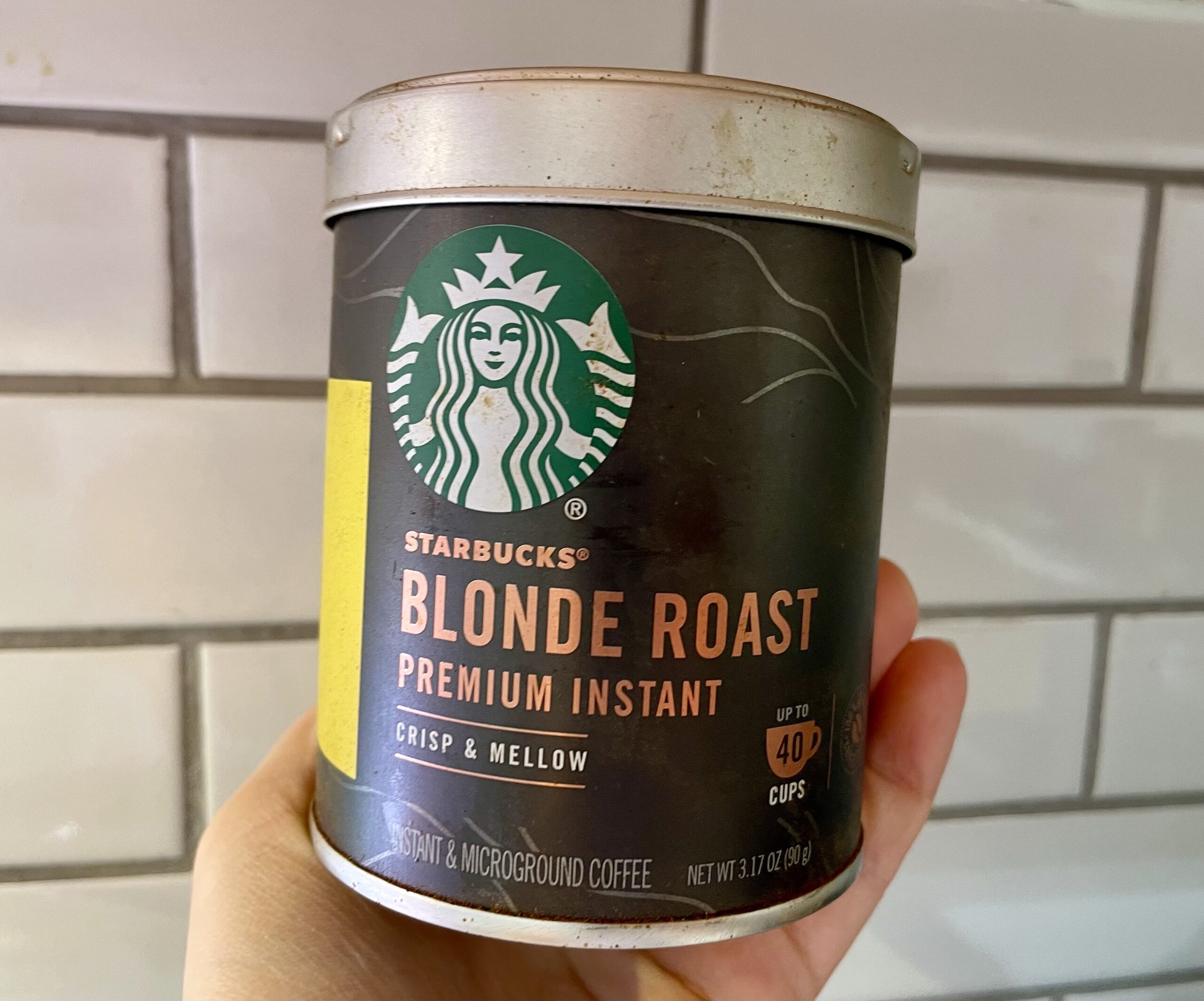 Hand holding a can of Starbucks Blonde Roast Instant Coffee against a white tile background
