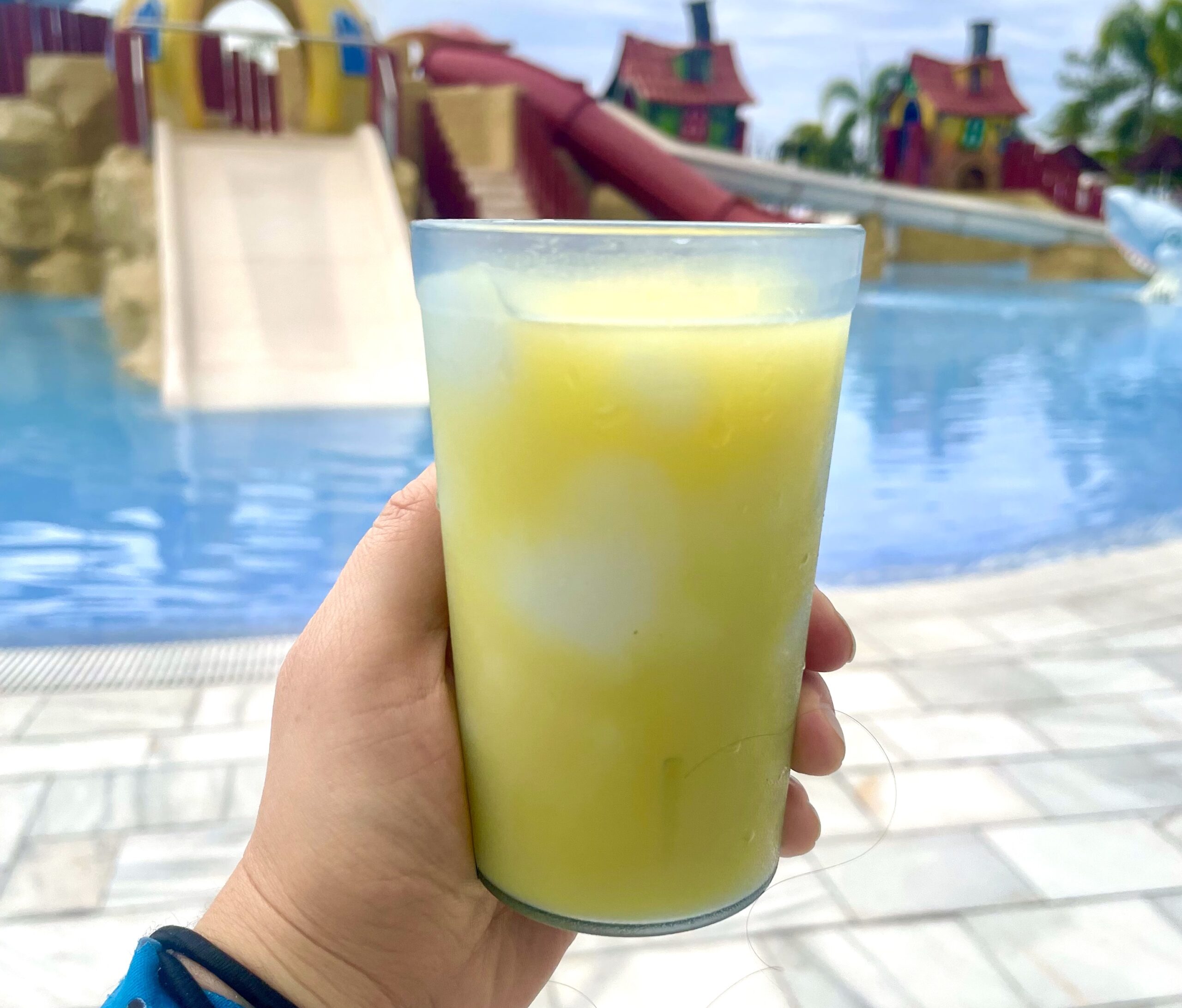 Hand holding a small plastic cup filled with pina colada and orange juice in Jamaica in front of a children's water slide area.